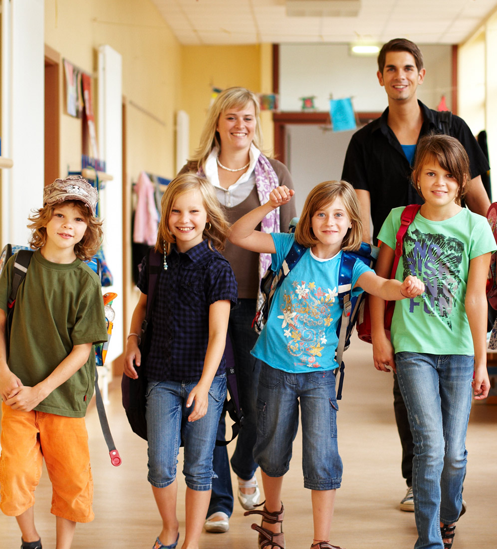 Symbolbild: Grundschülerinnen und Grundschüler im Schulflur, hinter ihnen Lehrkraft und Assistenzkraft