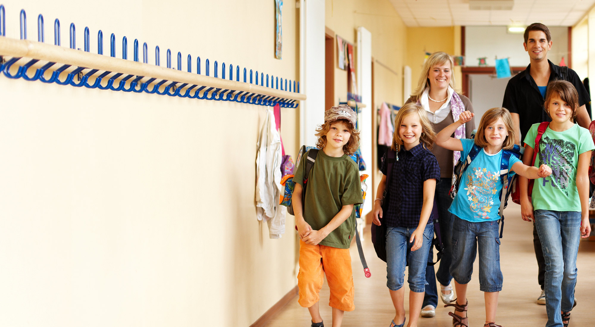 Symbolbild: Grundschülerinnen und Grundschüler im Schulflur, hinter ihnen Lehrkraft und Assistenzkraft