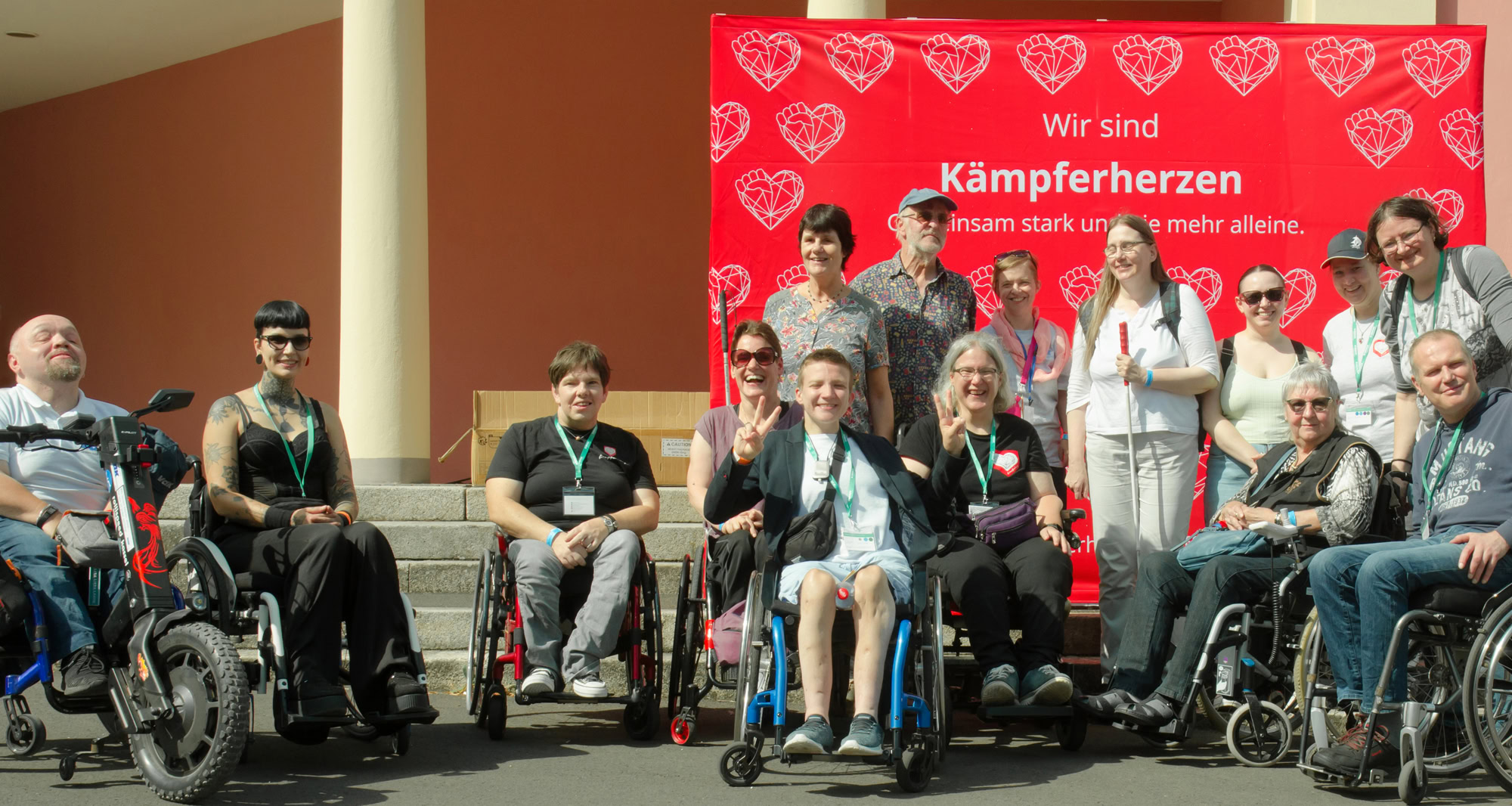 15 Personen mit und ohne Behinderung aus dem Fab-Umfeld stehen vor einer Fotowand, auf der zu lesen ist: „Wir sind Kämpferherzen. Gemeinsam stark und nie mehr alleine“.